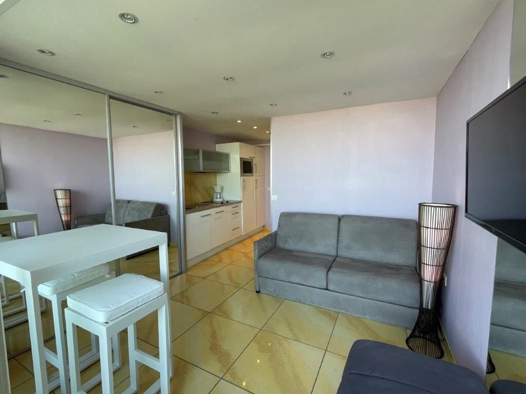 A modern living room with a sofa, a dining table with chairs, a TV, and a kitchen in the background.