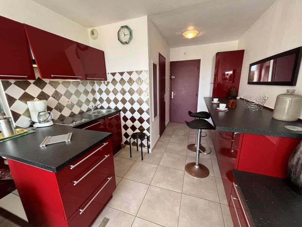 A kitchen with red cabinets, a black countertop, and a tiled backsplash. There are two stools and a coffee maker on the counter.