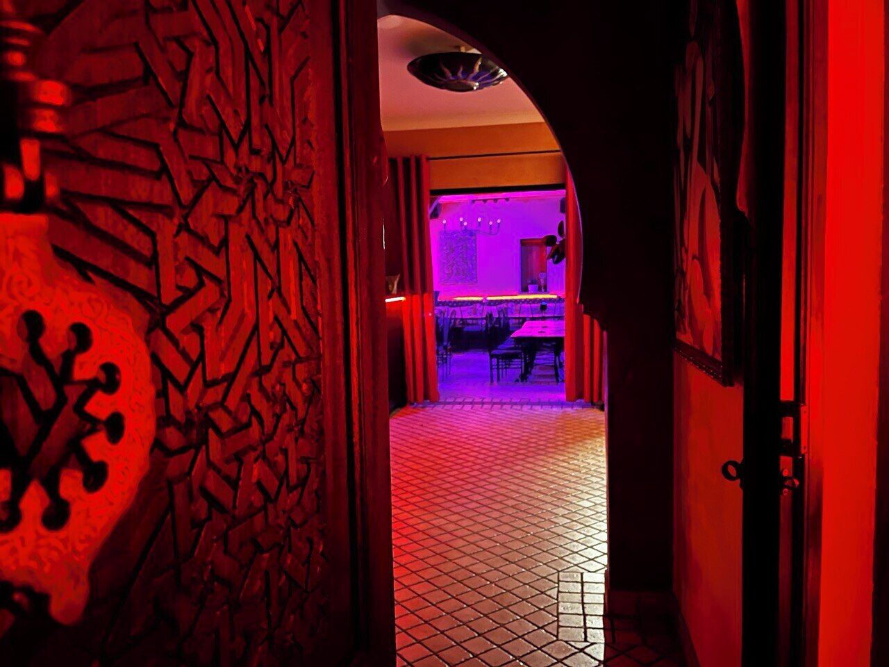 A hallway with a patterned door, a tiled floor, and a room in the background with a table and chairs.