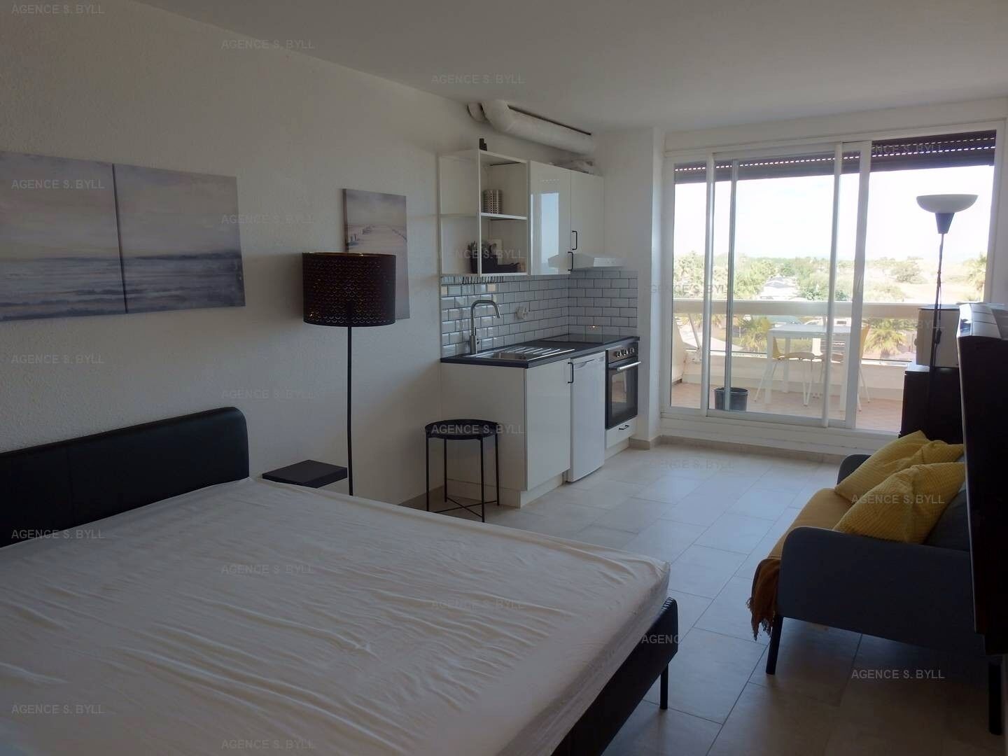 A modern bedroom with a large bed, a small kitchenette, and a balcony with a view.