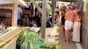 An outdoor covered area with a wooden structure, a table with a plant, and a crowd of people.
