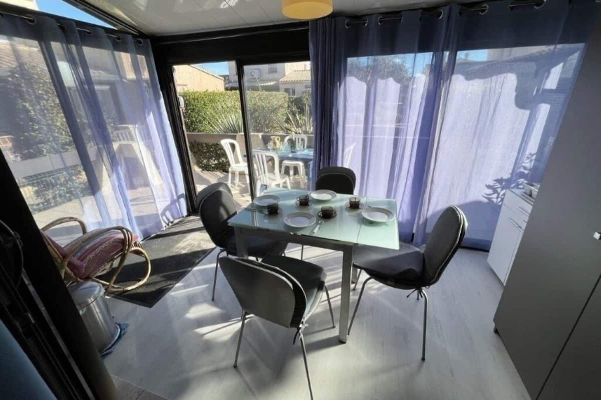 Un coin repas avec une table dressée pour quatre personnes, entourée de chaises, et de grandes fenêtres avec des rideaux.