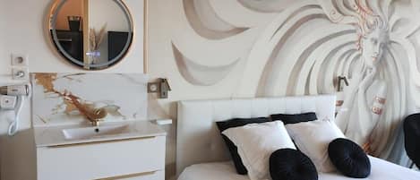 Un dormitorio moderno con una cama blanca, almohadas negras y blancas, un espejo redondo y un mural decorativo en la pared.