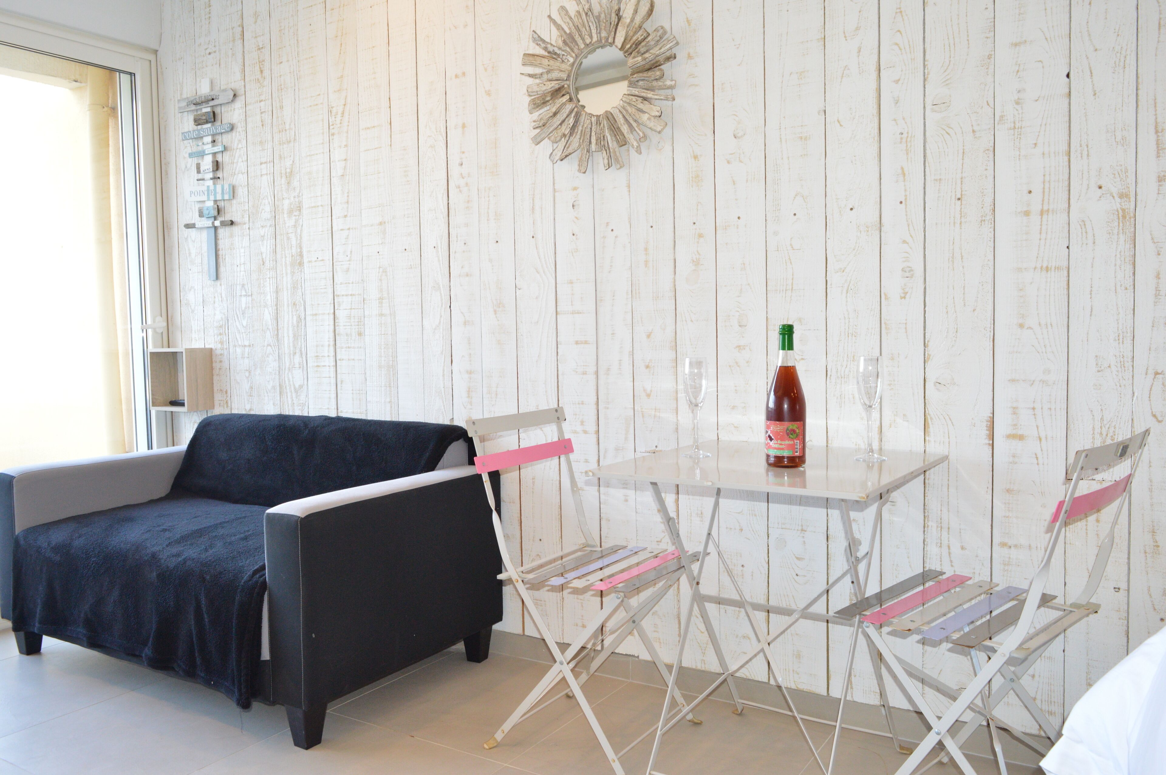 Une pièce avec un mur en bois peint en blanc, un canapé noir, une petite table sur laquelle se trouvent une bouteille et un verre, et une chaise pliante.