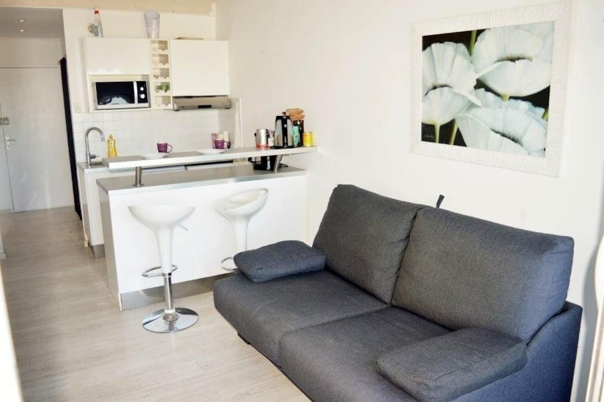 Une cuisine moderne avec des armoires blanches, un plan de travail blanc et un tabouret de bar. On aperçoit un canapé gris au premier plan et un tableau représentant des fleurs blanches accroché au mur.