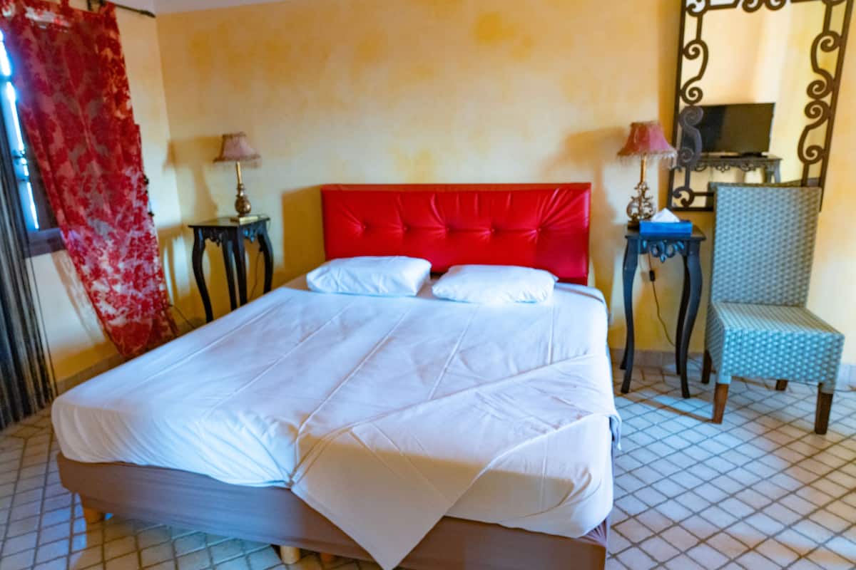 Un lit avec une tête de lit rouge, deux tables de chevet avec des lampes, une chaise et un téléviseur posé sur un meuble.