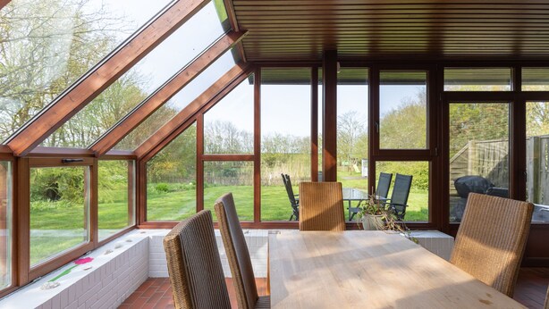 A sunlit patio with a wooden table and wicker chairs, large windows, and a view of a garden. - Vacation home Haus am Deich in Reußenköge for up to 6 people (Reußenköge)
