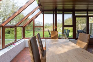Een zonnig terras met een houten tafel en rieten stoelen, grote ramen en uitzicht op een tuin.