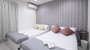 Ein Hotelzimmer mit zwei Betten, grauer Bettwäsche, gemustertem Wandpapier, einer Klimaanlage, einem kleinen Tisch mit einem Buch und einem Mülleimer.