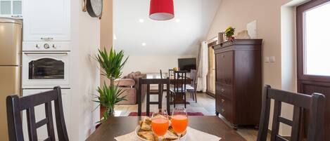 A dining area with a wooden table, two chairs, a bowl of food, and two glasses of orange juice. There is a red pendant light above the table, a clock on the wall, and a plant in the corner.