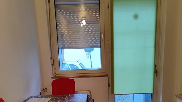 Ruokailutila, jossa on kuvioidulla pöytäliinalla peitetty pöytä, kaksi punaista tuolia, ikkuna, jossa on sälekaihtimet, ja ovi, joka johtaa ulos.