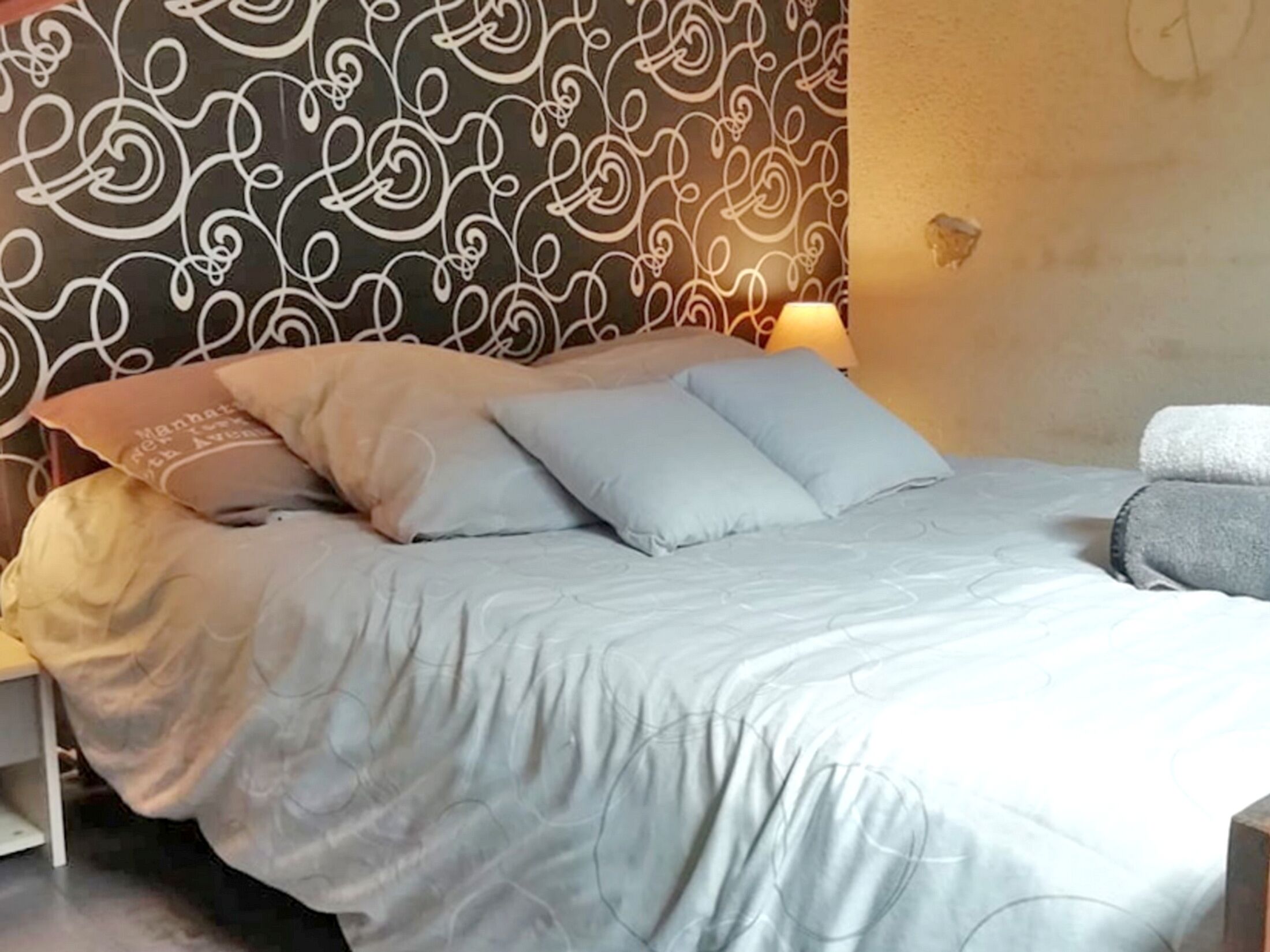 Una cama bien hecha con una colcha azul claro y almohadas, una mesita de noche con una lámpara, y una pared con un diseño ornamental de volutas.