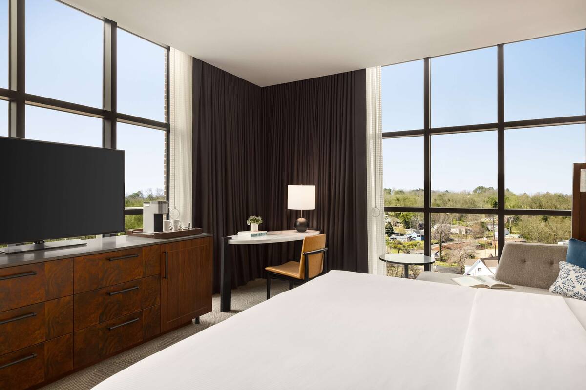 A hotel room with a large bed, a desk with a chair, a TV, and a view of the outside.