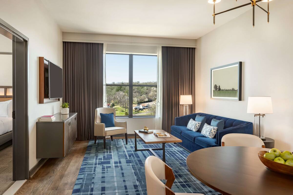A hotel room with a large window, a blue sofa, a wooden coffee table, a dining table, and a TV on a cabinet.