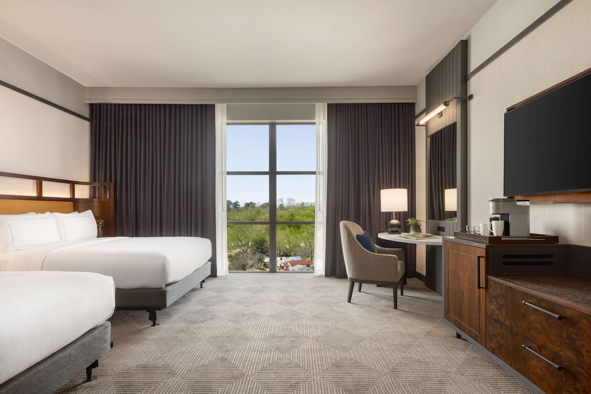 A hotel room with two beds, a desk with a chair, a television, and a large window with curtains.