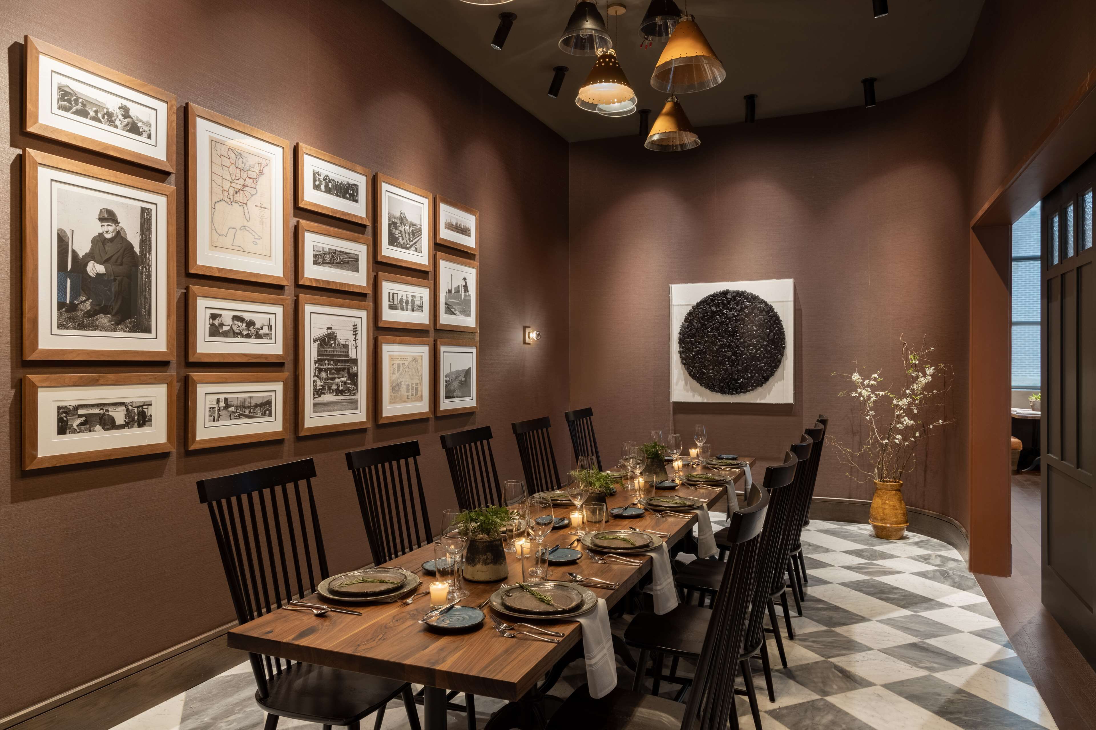 A dining area with a long wooden table set for six, surrounded by black chairs. The walls are adorned with framed black and white photographs.