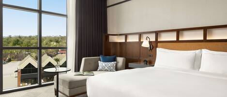 A hotel room with a large bed, a small round table, a chair, a nightstand, and a large window offering a view of buildings and trees.