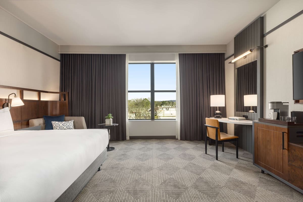 A hotel room with a large bed, a desk with a chair, a small table with a plant, and a view of the outdoors through a window.