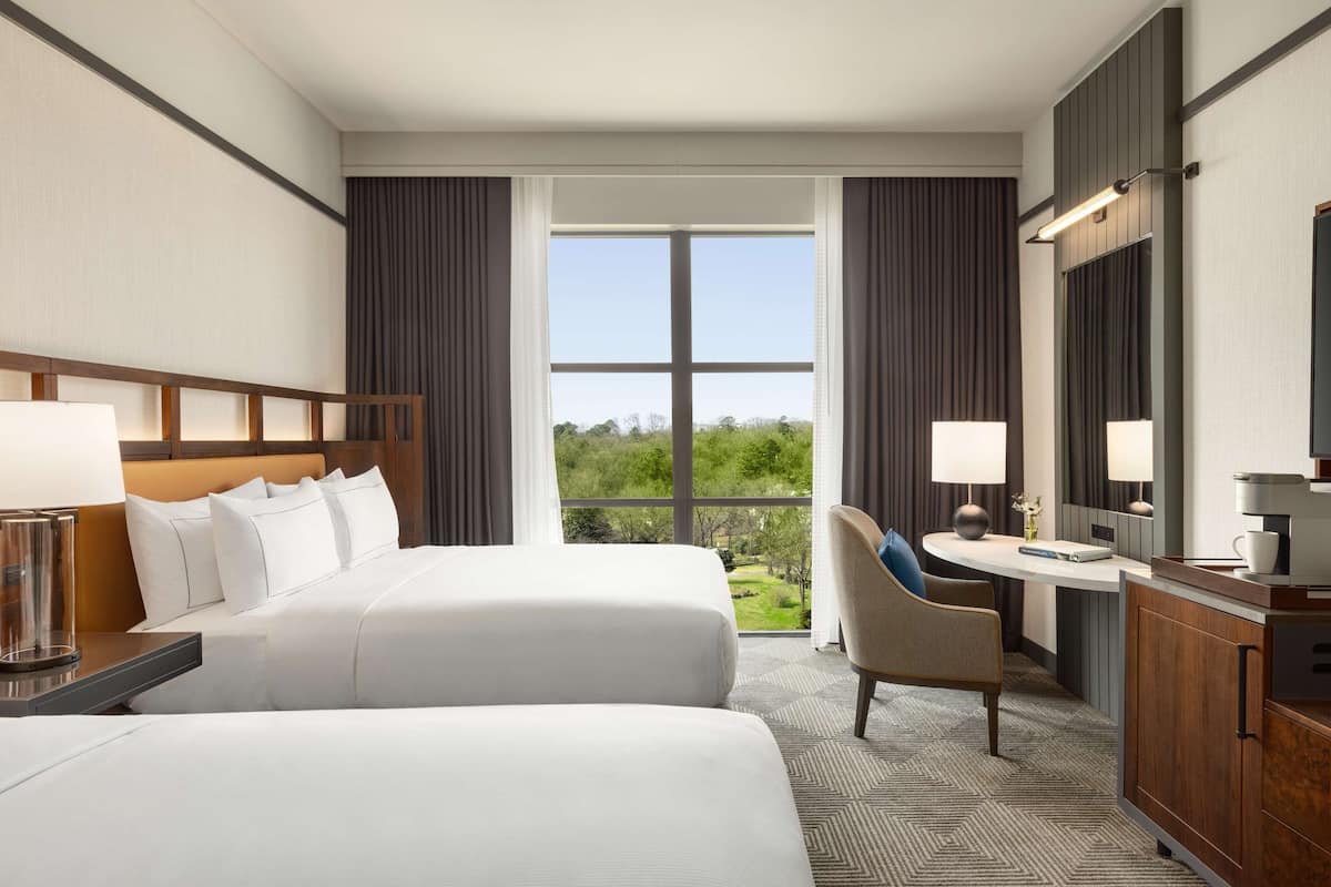 A hotel room with two beds, a desk, a chair, a coffee machine, and a large window with a view of greenery.