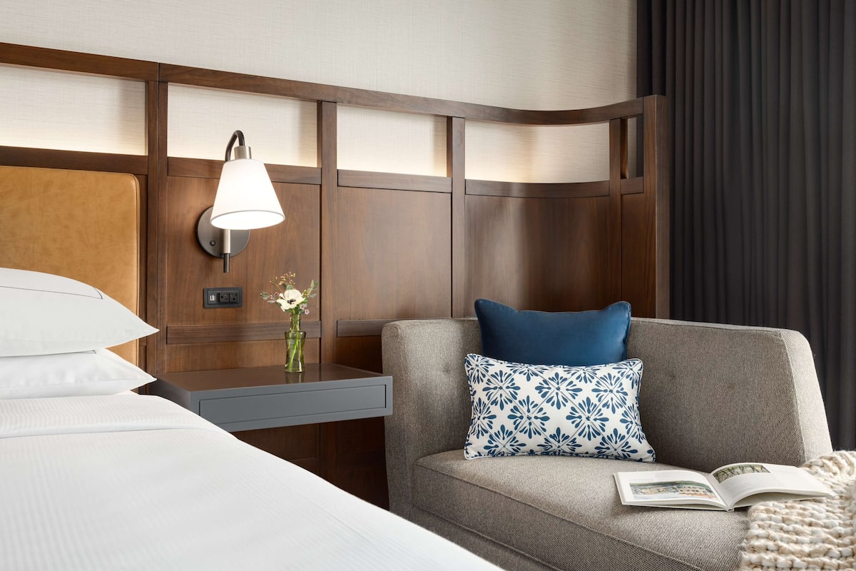 A hotel room with a bed, a nightstand, a lamp, a sofa with a pillow, and a book.