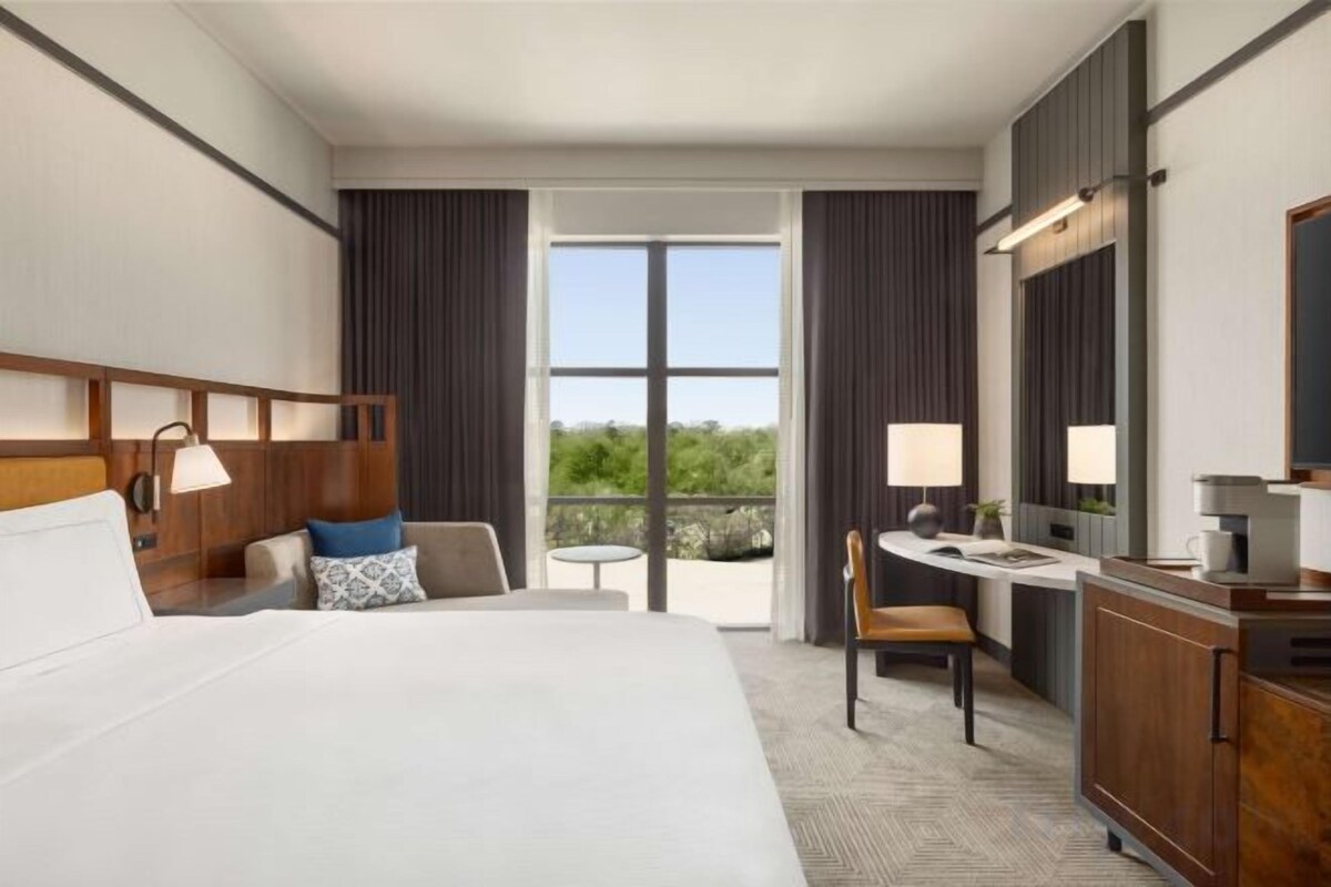 A hotel room with a large bed, a desk, a chair, a small table, and a balcony with a view.