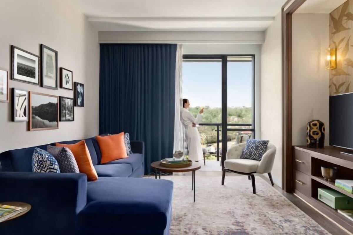 A modern hotel room with a large window, a sofa, a chair, a TV, and framed pictures on the wall.