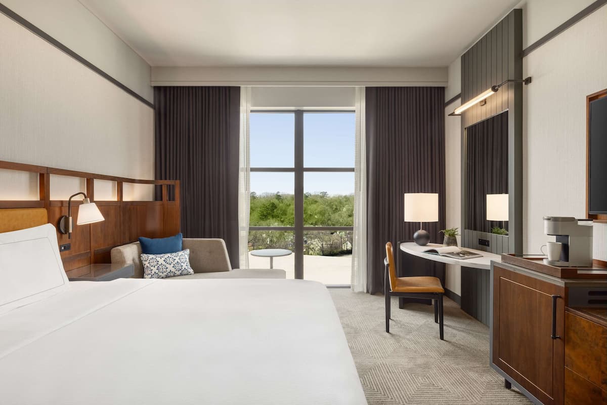 A hotel room with a large bed, a desk, a chair, a small table, and a view of the outdoors.