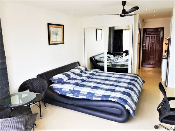 A modern bedroom with a large bed, a small round table, and a chair. There is a ceiling fan and a door leading to another room. - 11th Floor Studio at View Talay 1 (Pattaya)