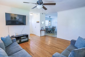 A modern living room with a flat-screen TV, grey sofas, wooden flooring, a ceiling fan, and a dining area with a table and chairs.