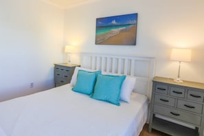 A bedroom with a bed, two nightstands, a painting of a beach, and a white headboard.