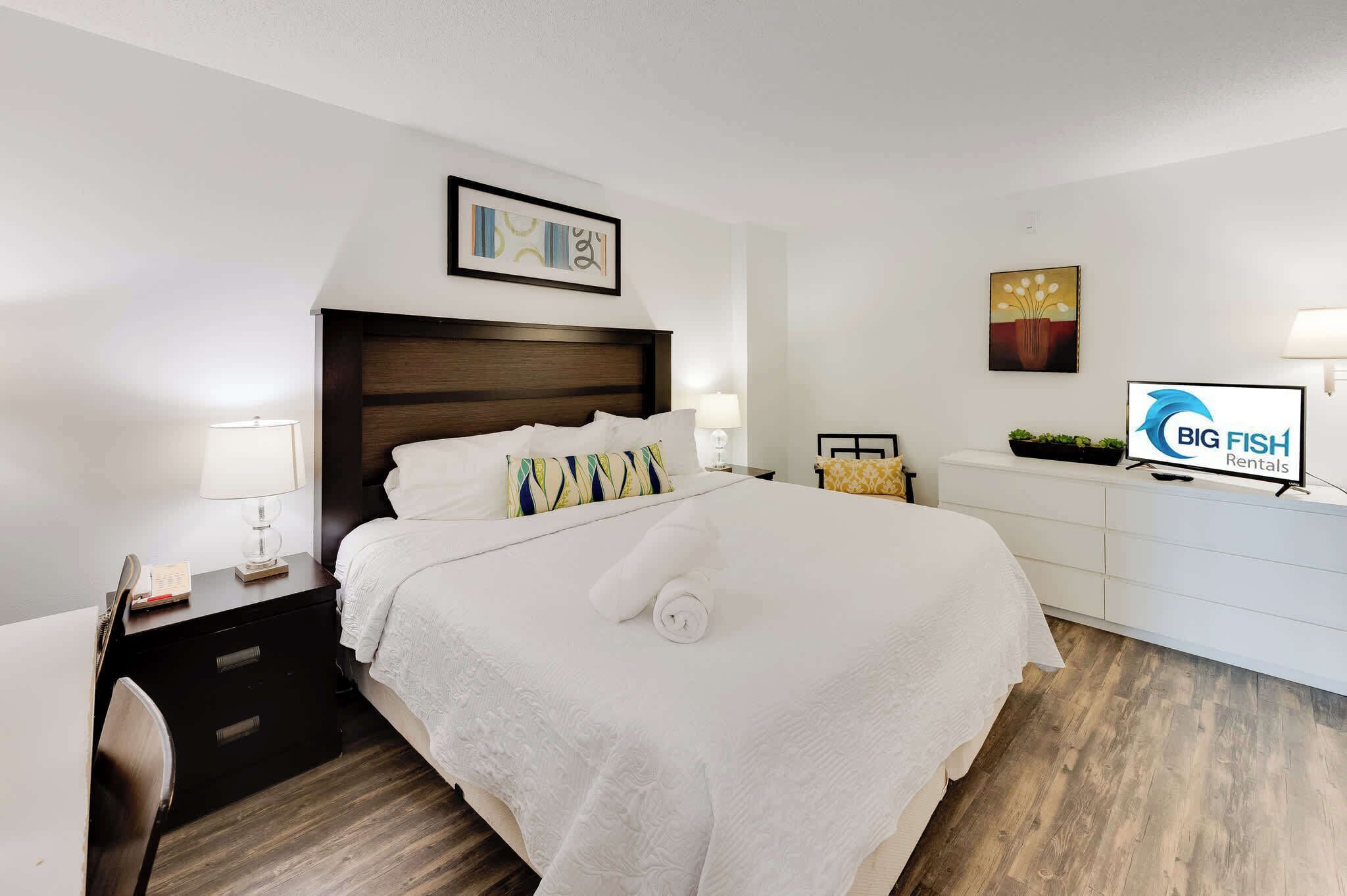 A hotel room with a large bed, a desk with a lamp, a chair, a TV, and artwork on the walls.