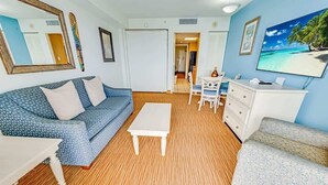 A living room with a blue sofa, a white coffee table, a white dresser, and a television on the wall. - Oceanfront,Heated Pools,Views New, Penthouse Level (Myrtle Beach)