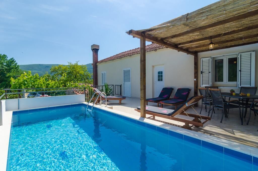 Una piscina con sillas de descanso y una mesa, rodeada por una pérgola de madera y un edificio con contraventanas.
