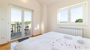 Un dormitorio con una cama grande, un ventanal corredero que da a un espacio exterior con sillas y una ventana con vista a la vegetación.