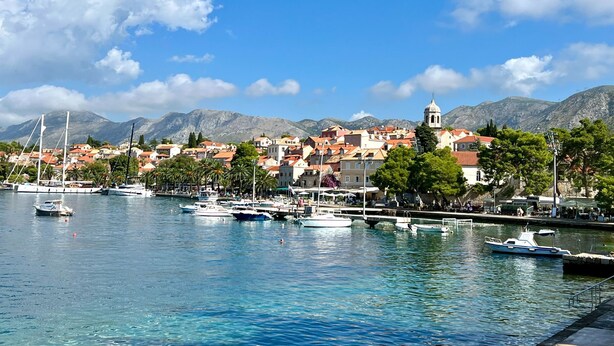 Un pueblo costero con un puerto deportivo, edificios con techos rojos y una montaña de fondo.