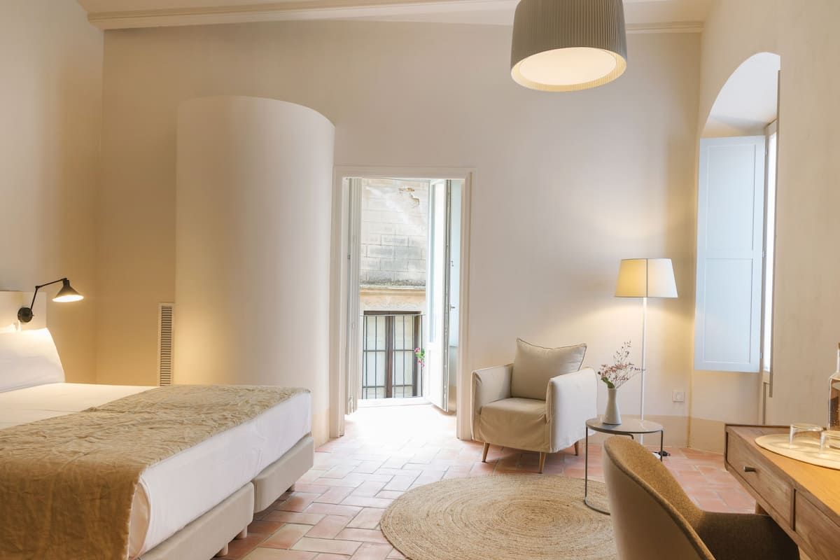 A hotel room with a large bed, a sitting area with a chair and table, a desk with a lamp, and a large mirror.