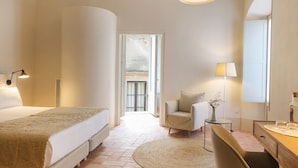 A hotel room with a large bed, a sitting area with a chair and table, a desk with a lamp, and a large mirror.