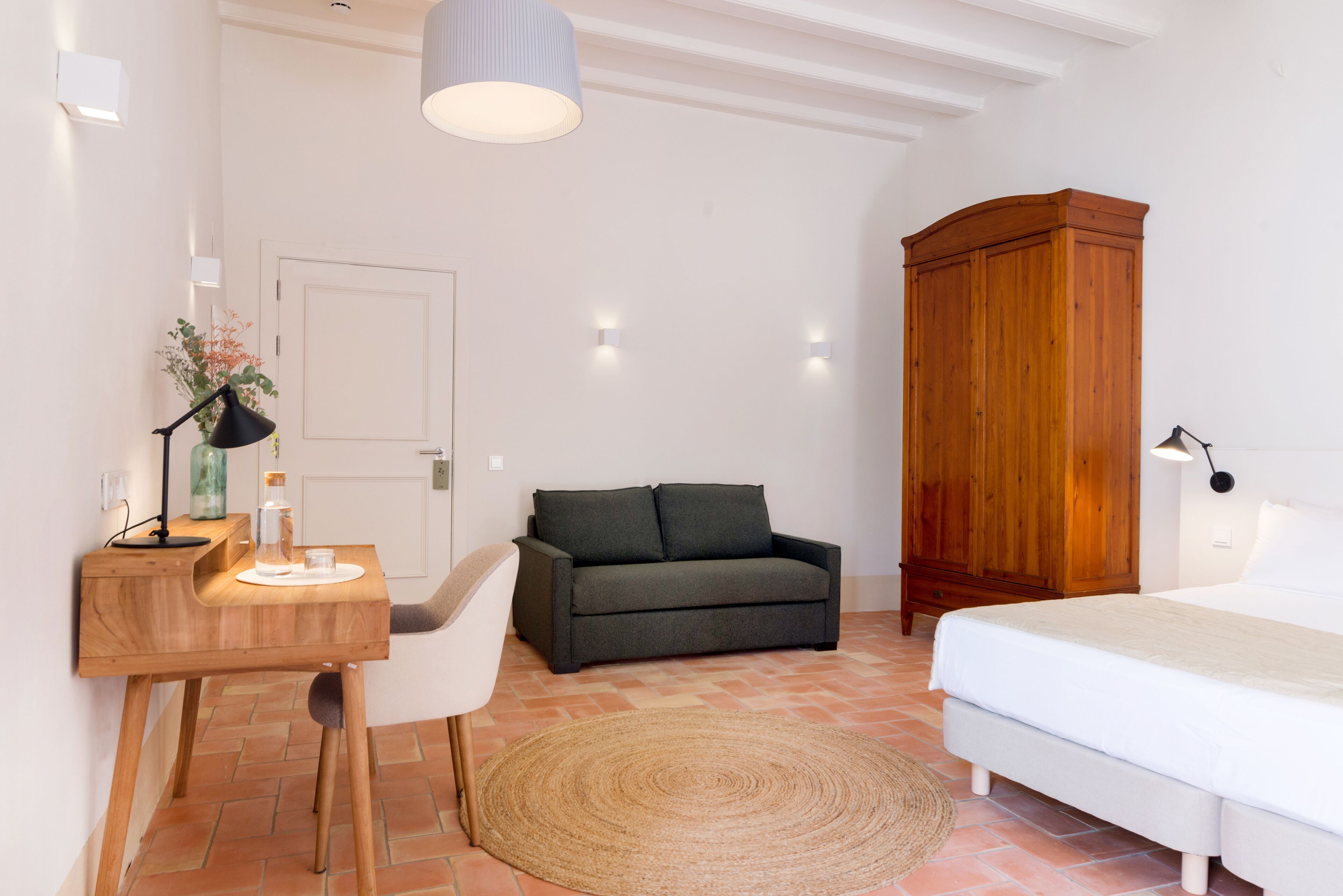 Een slaapkamer met een bed, een bureau met stoel, een donkere bank, een houten kledingkast en een geweven vloerkleed.