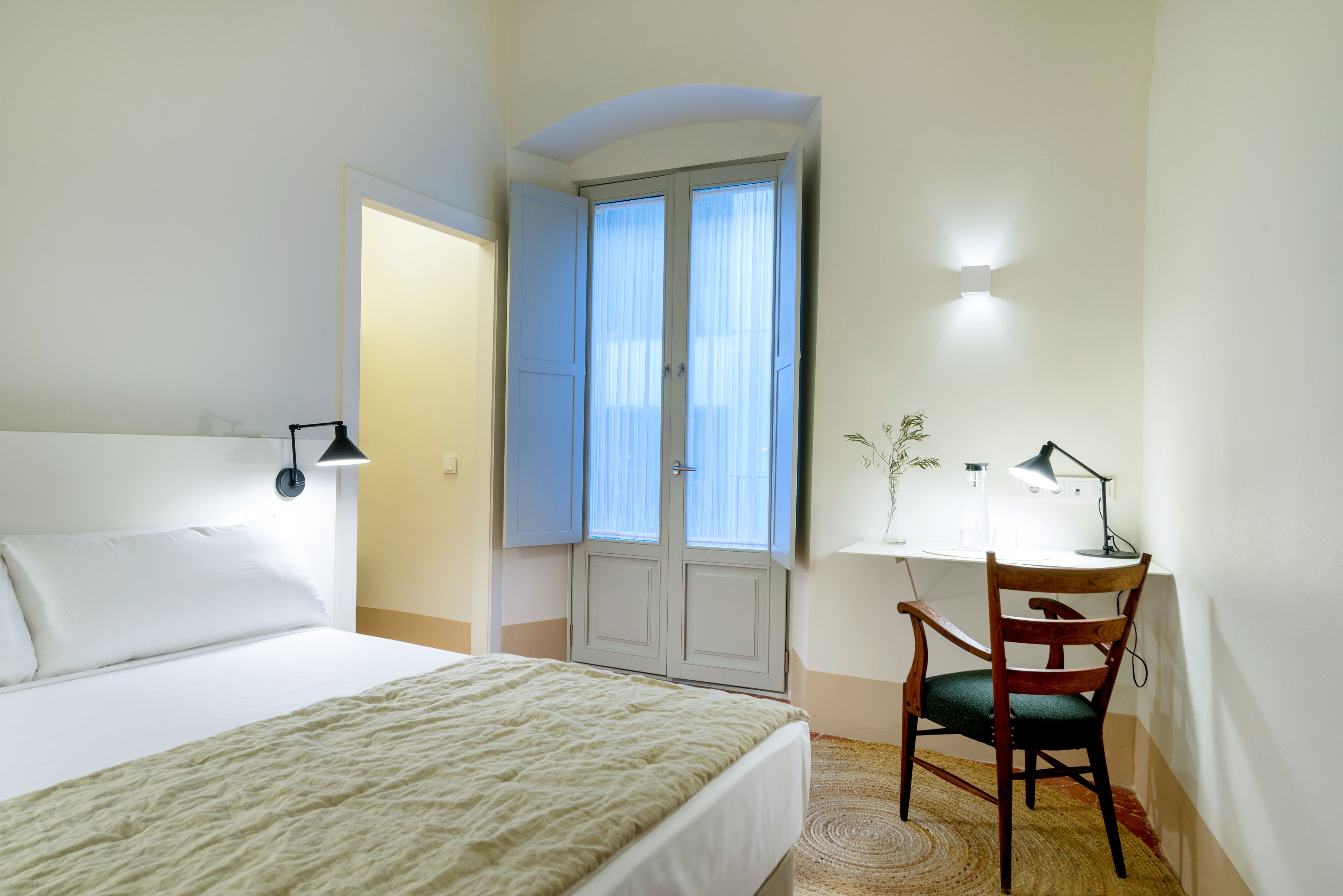 A bedroom with a bed, a desk with a chair, a window with curtains, and a wall-mounted light.
