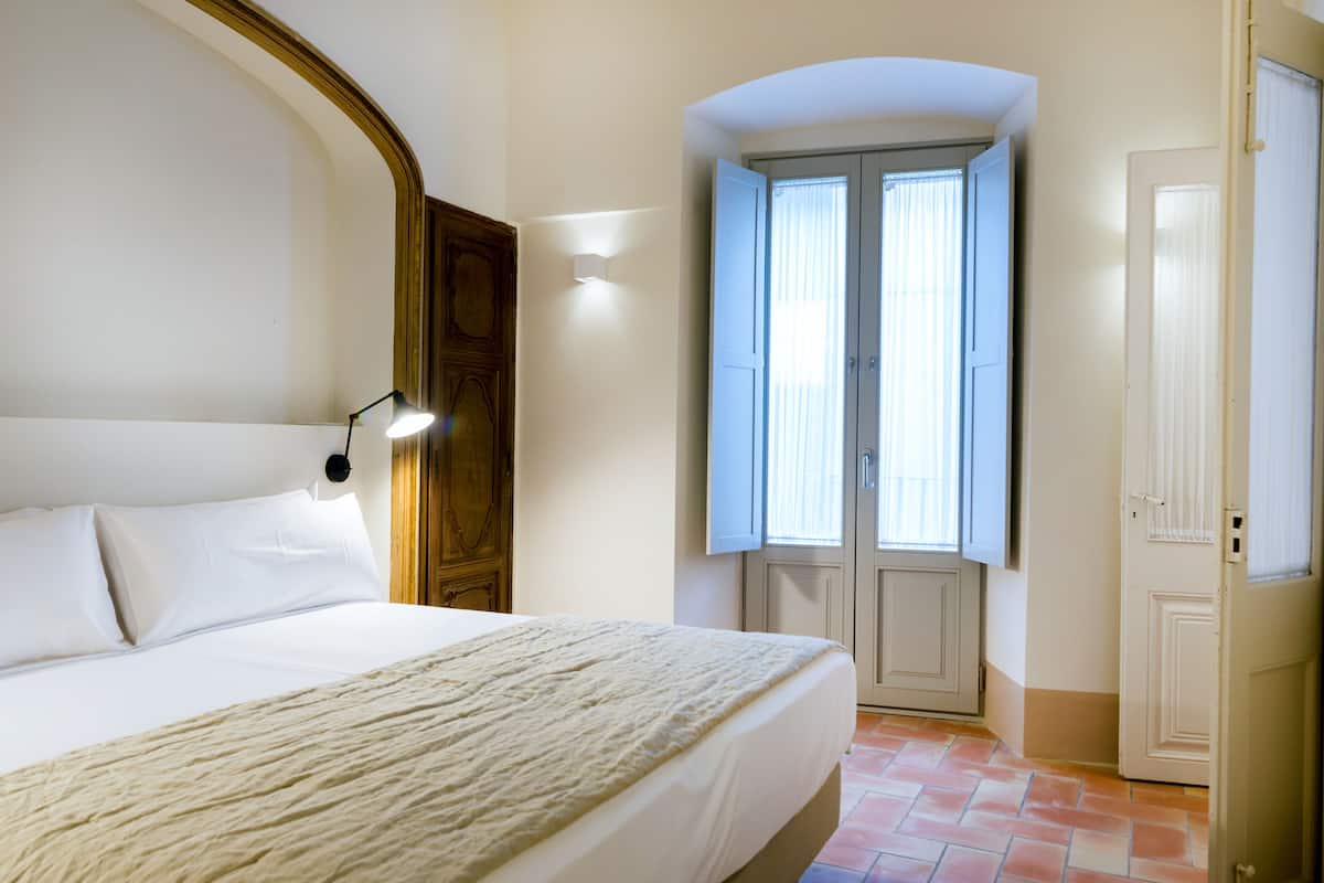 A bedroom with a large bed, a wooden wardrobe, a nightstand with a lamp, and a window with curtains.