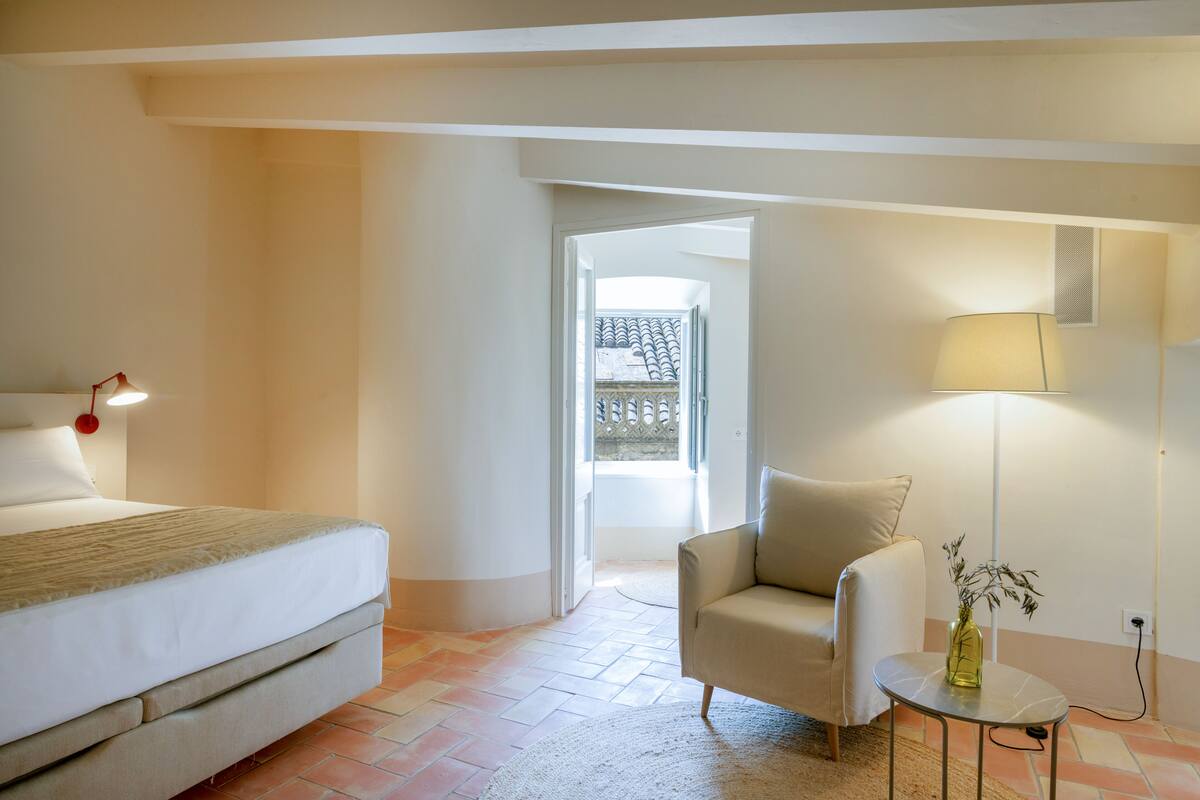 A bedroom with a bed, a chair, a small table with a lamp, and a vase with plants.