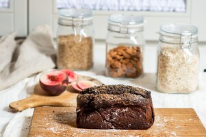 Una hogaza de pan sobre una tabla de madera con un rodillo, rodeada de frascos de cereales y media fruta.