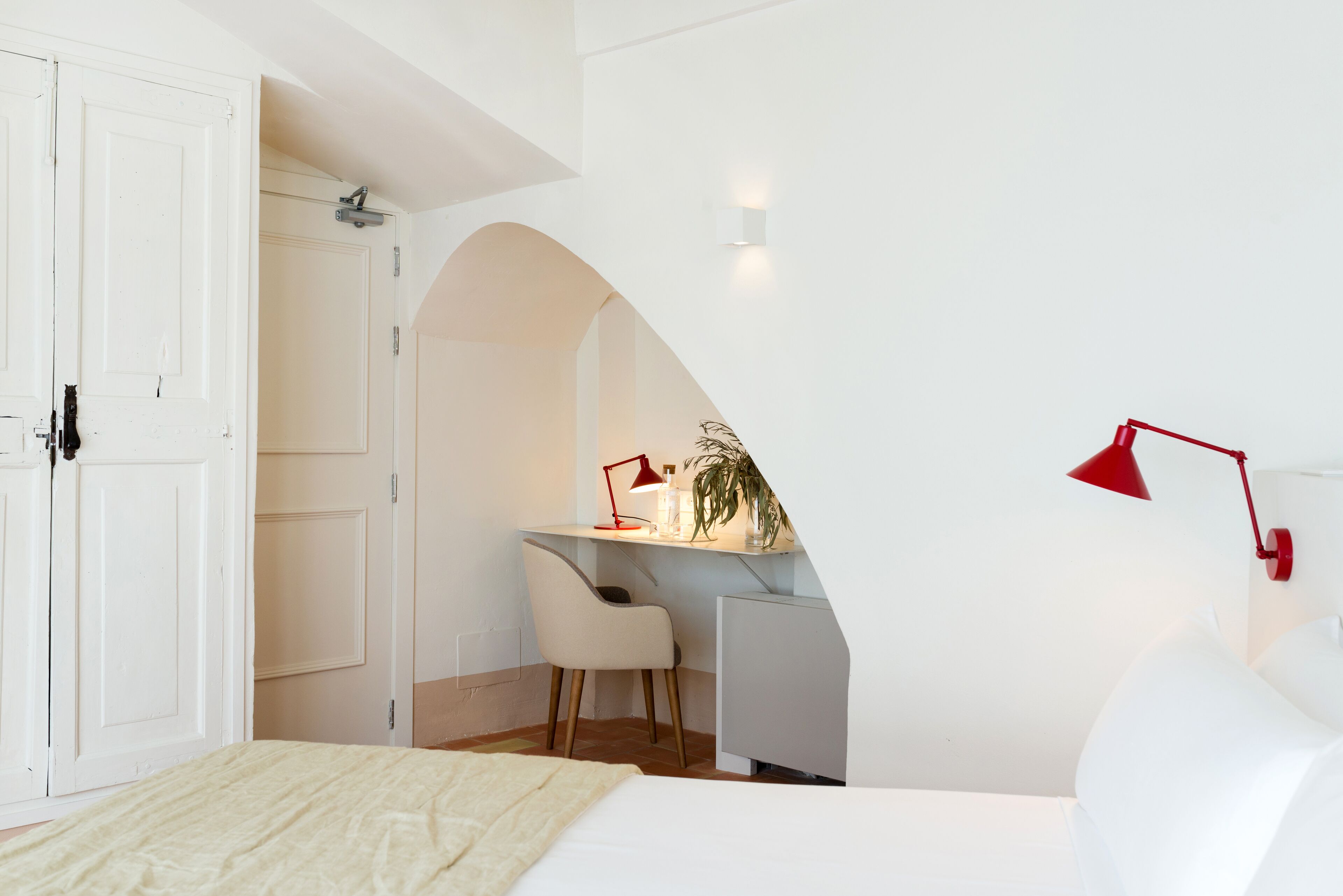 A bedroom with a bed, a desk, and a chair. There is a white door and a white wall with a red lamp.