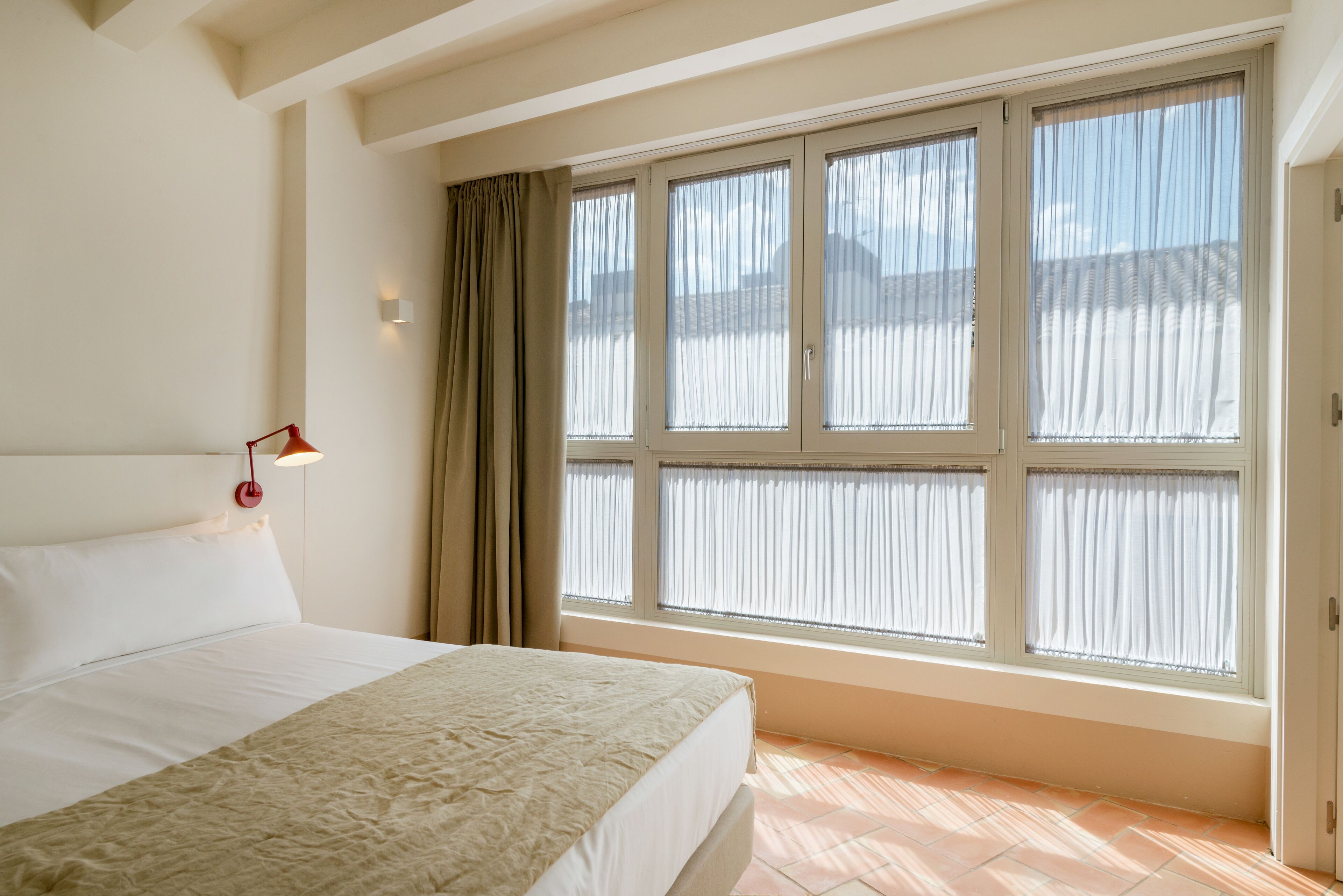 A hotel room with a large bed, a window with curtains, and a wall-mounted light.