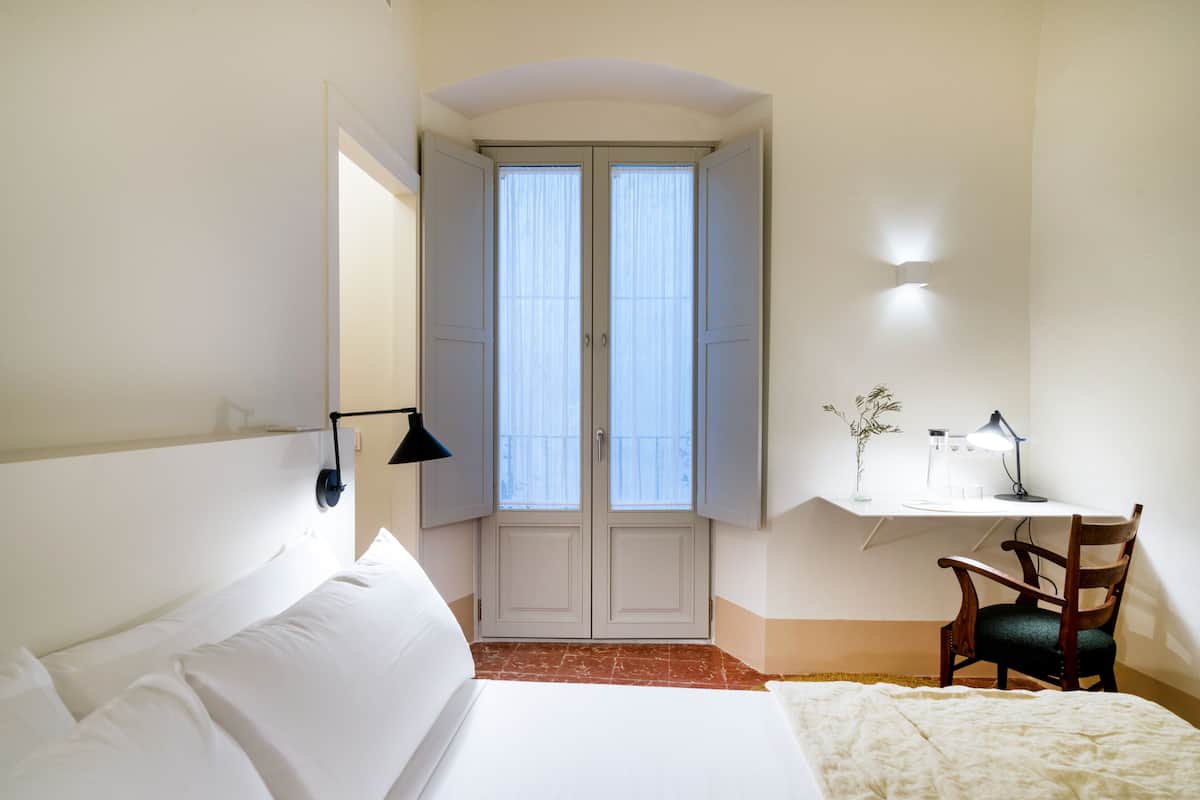 A hotel room with a bed, a desk with a chair, a window with curtains, and a wall-mounted lamp.