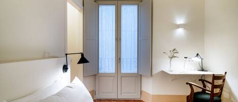 A hotel room with a bed, a desk with a chair, a window with curtains, and a wall-mounted lamp.