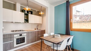 Una cucina moderna con mobili bianchi, un piano di lavoro in legno e un forno integrato. Si vede un tavolo da pranzo con sedie bianche e una finestra con vista sugli edifici.