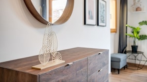 Un comò in legno con un elemento decorativo a forma di foglia sulla sommità, uno specchio rotondo e opere d'arte appese alla parete.