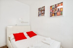 A hotel room with a bed, white bedding, red pillows, a white headboard, a bedside table with a lamp, and two framed pictures on the wall.