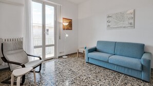 A modern living room with a blue sofa, a chair, a small table, and a floor lamp. There is a large window with curtains and a door leading outside. - Lulli Cozy Apartment with Balcony (Florence)